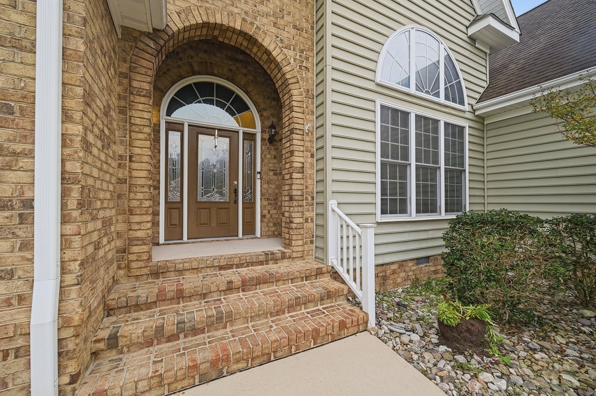 Brick home entrance with arched doorway and sidelights leading to white wooden porch, concrete walkway, and green landscaping.