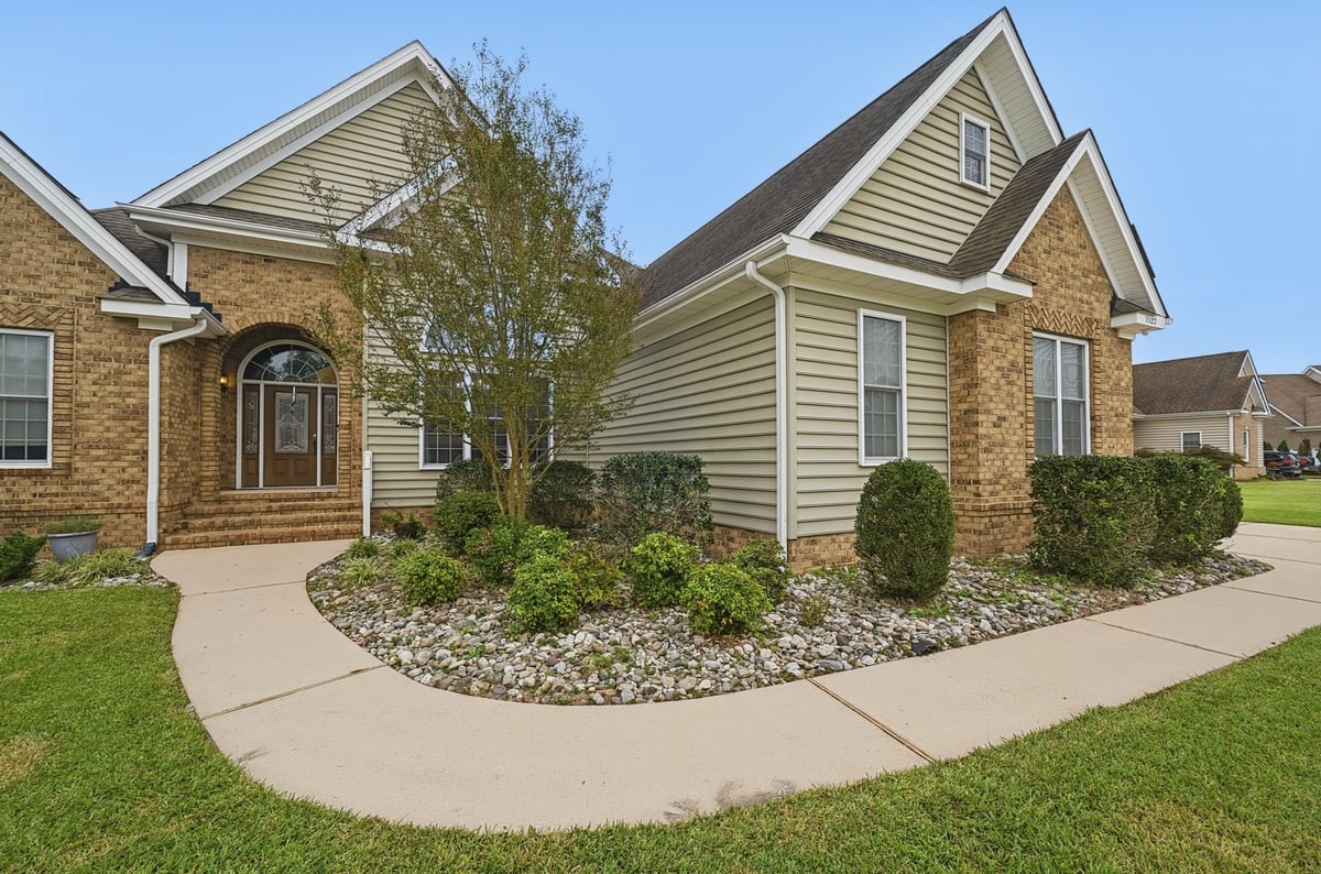 Two-story brick and siding suburban house with manicured front lawn, landscaping, and concrete driveway on a clear sunny day.