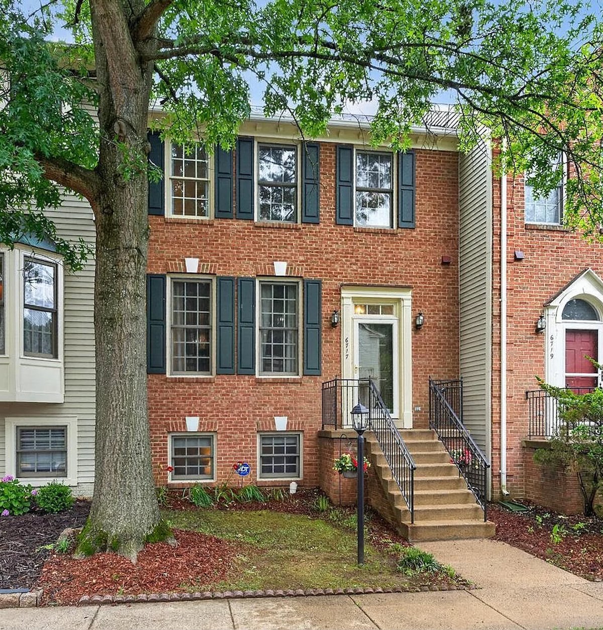 Red brick townhouse with black shutters, front steps, and large tree in yard